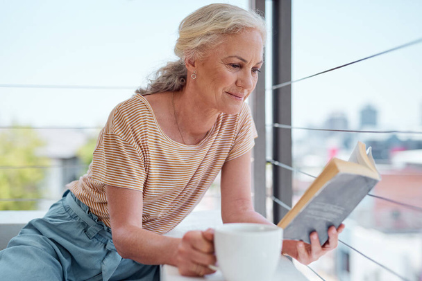 Tantos libros y un montón de tiempo. una mujer madura leyendo un libro y tomando café en su balcón en casa - Foto, imagen
