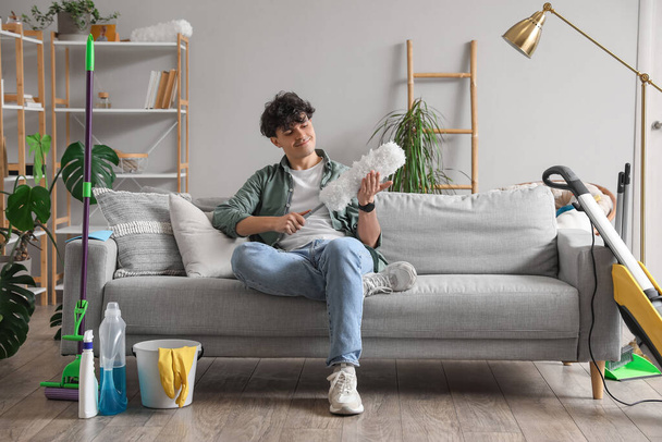 Junger Mann mit Staubwedel sitzt zu Hause auf Sofa - Foto, Bild