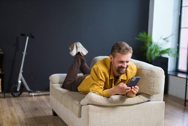 Smiling man enjoying time at home, sitting on sofa with phone. He appears handsome and content engages in texting or messaging on mobile device. High quality photo - Fotó, kép