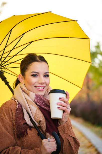Valkoihoinen nuori nainen kahvin ja keltaisen sateenvarjon kanssa hymyilee ja katsoo kameraa syksyllä. - Valokuva, kuva