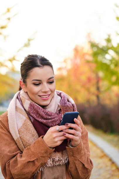 Kaukaska młoda kobieta korzystająca z telefonu uśmiechnięta jesienią. - Zdjęcie, obraz