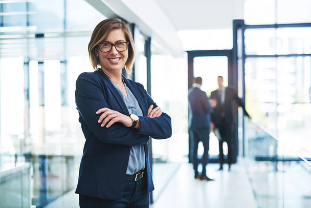 Being in this office is everything I dreamed of. Cropped portrait of an attractive young businesswoman standing with her arms folded in an office with her colleagues in the background - Foto, immagini