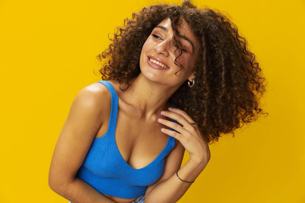 Mavi tişörtlü kıvırcık saçlı kadın. Elleriyle sarı arkaplan işaretleri, kameraya bak, dişleriyle ve mutlulukla gülümse, kopyalama alanı. Yüksek kalite fotoğraf - Fotoğraf, Görsel