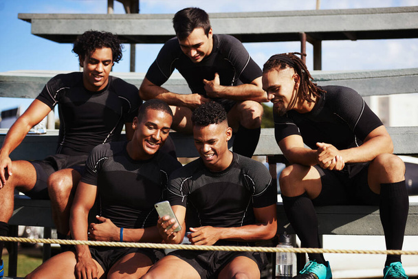 Las redes sociales están zumbando con la temporada de rugby acumulada. un joven usando un smartphone en un partido de rugby rodeado de sus compañeros de equipo - Foto, imagen