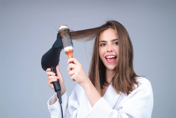 Woman in bathrobe combing hair, drying hairs with hairdryer. Portrait of female model with a comb brushing hair. Girl with hair brush and blow dryer. Hair care and beauty. Morning routine - Fotografie, Obrázek