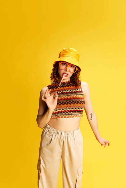 Portrait of lovely young girl with curly hair, posing in panama, glasses and knitted top, eating bubble gum over yellow studio background. Concept of human emotions, youth culture, fashion, lifestyle - 写真・画像