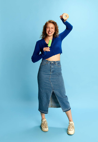 Full-length portrait of happy, emotional young girl in blue top and jeans skirt holding ice cream and donut against blue studio background. Concept of human emotions, youth culture, fashion, lifestyle - Foto, afbeelding