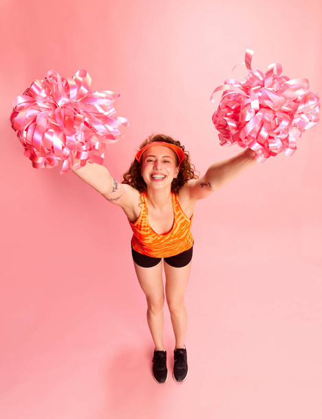 Full-length portrait of young girl, cheerleader in sportswear posing in motion against pink studio background. Concept of human emotions, youth culture, fashion, sportive lifestyle - Fotoğraf, Görsel