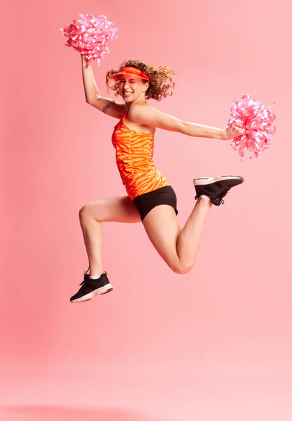 Full-length portrait of young girl, cheerleader in sportswear posing in motion against pink studio background. Concept of human emotions, youth culture, fashion, sportive lifestyle - Zdjęcie, obraz