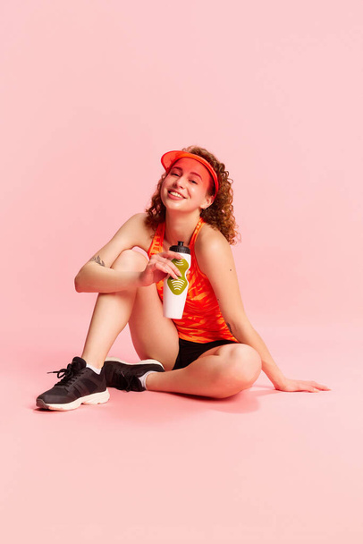 Portrait of young smiling girl in sportswear sitting on floor and drinking water after training against pink studio background. Concept of human emotions, youth culture, fashion, sportive lifestyle - Valokuva, kuva