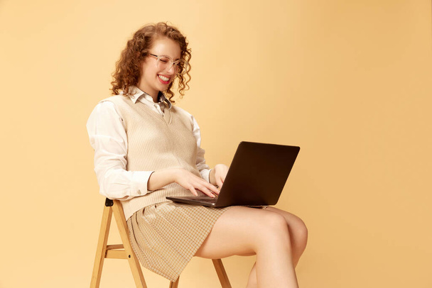 Portrait of young beautiful girl in formal clothes sitting on chair and having online video call on laptop over studio background. Human emotions, youth culture, fashion, lifestyle, business concept - 写真・画像