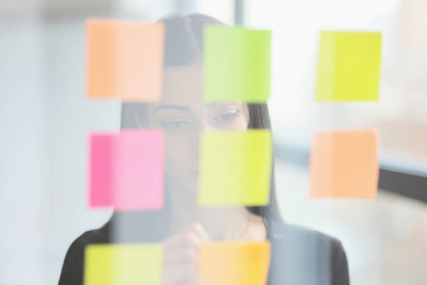 Business lady reads notes on memory sticks hanging on glass wall in office. Female employee plans work tasks in coworking. Marks on color papers - Foto, imagen
