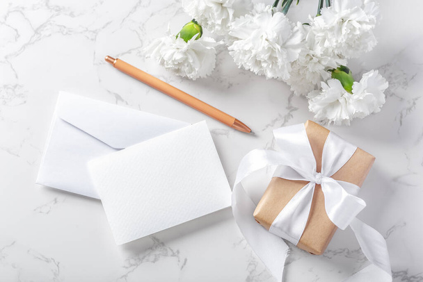 White paper card and envelope with gift box and flowers on marble background, top view - Foto, immagini