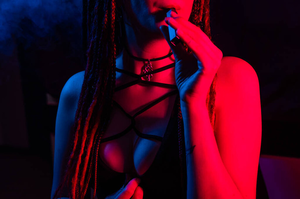 Caucasian girl with dreadlocks smokes a vape in red blue light - Foto, afbeelding
