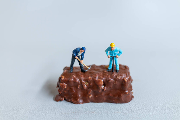 ミニチュア人、従業員は灰色の背景にチョコレートバーを作っています。世界チョコレートデーのコンセプト - 写真・画像