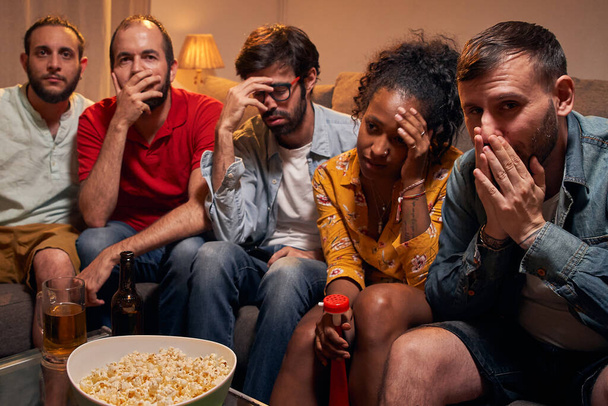Sad and disappointed group of multiracial friends watching soccer game on TV. People gathered at home in evening to watch their sport team lose game. Negative feelings and expressions. - Photo, image