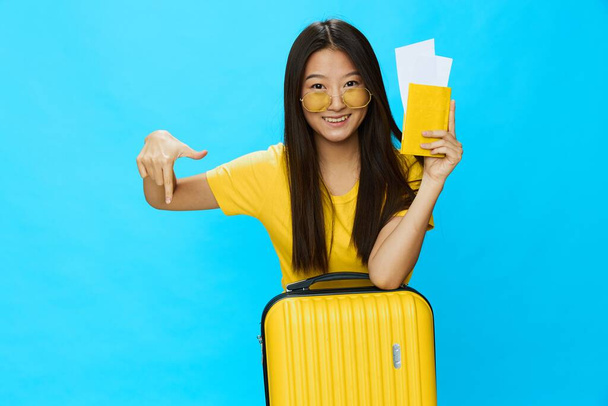 Ασιάτισσα που ταξιδεύει με κίτρινη βαλίτσα και εισιτήρια με διαβατήριο στο χέρι, τουρίστρια που ταξιδεύει με αεροπλάνο και τρένο με αποσκευές σε μπλε φόντο. Υψηλής ποιότητας φωτογραφία - Φωτογραφία, εικόνα
