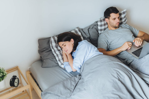 Pretty female sleeps in bed, sees pleasant dreams, while her husband works on laptop computer, prepares report, tries not to interrupt his wife sleep. Family couple in bedroom. Rest concept. - Photo, Image