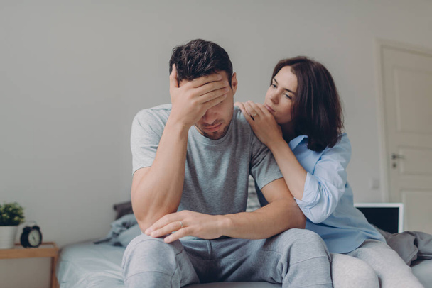 Attractive female consoles her sad boyfriend who has depression and some problems, pose at bedroom on bed, sit against domestic interior. Wife and husband try solve difficult situation in life. - Foto, imagen