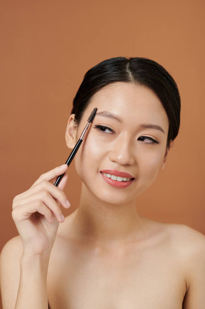Studio portrait of smiling Vietnamese young woman brushing her eyebrows - Photo, Image