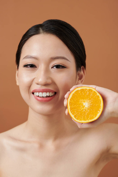 Portret uśmiechniętej młodej kobiety z nieskazitelną skórą trzymającej połowę pomarańczy - Zdjęcie, obraz