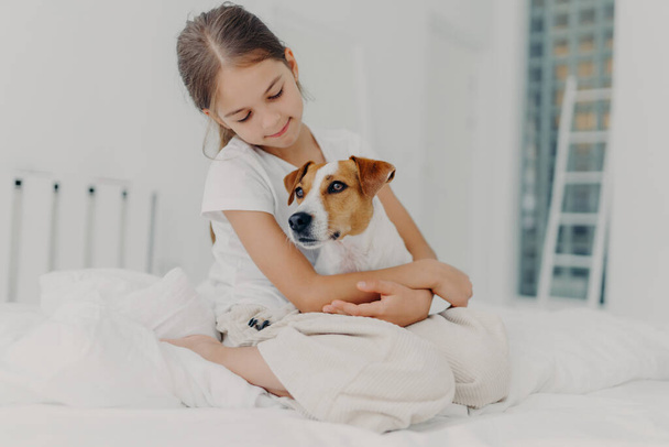 Photo of relaxed beautiful small girl plays with pedigree dog, embraces favourite animal, dressed in casual pyjamas, sits on bed, expresses love to pet, poses in light bedroom. Happy childhood concept - Fotó, kép