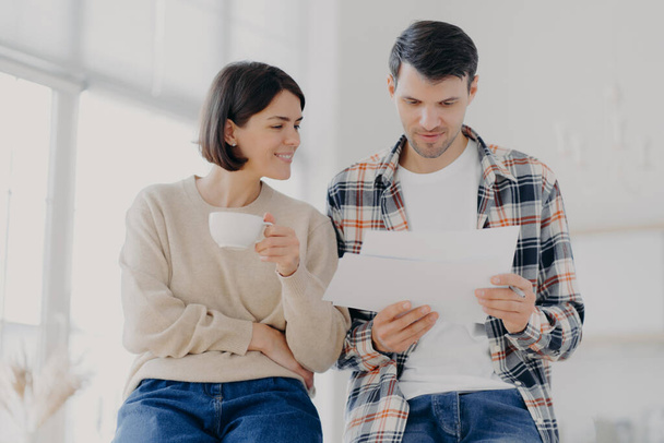 Serious couple study documents together, have serious looks, drink coffee, dressed in casual wear, plan their budget, pose in spacious light room, do paperwork, busy preparing financial report - Photo, Image