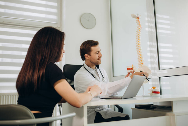 脊椎モデルの脊椎への患者のポイントに話す女性医師治療の原理を伝える。医務室のテーブルに座っている若い女性に真剣な医者が話している。脊椎動物学の概念. - 写真・画像