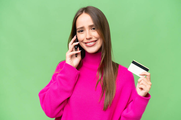 Young beautiful caucasian woman over isolated background keeping a conversation with the mobile phone and holding a credit card - Foto, Bild