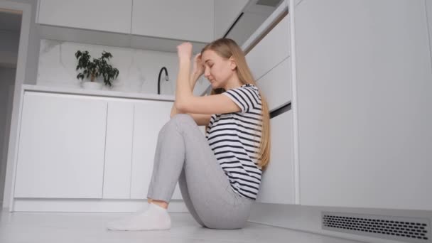 Biała kobieta cierpiąca na silny ból głowy, siedząca na podłodze w kuchni, czująca się wyjątkowo chora.. - Materiał filmowy, wideo