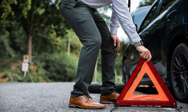 Asian businessman stranded with broken car and red warning triangle on the side of the road. Waiting for roadside assistance and repair services. - Foto, imagen