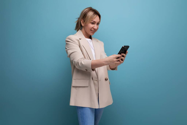 blond jong vrouw in een jasje maakt een betaling met behulp van een telefoon op een studio achtergrond. - Foto, afbeelding