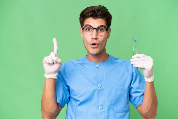 Joven dentista sosteniendo herramientas sobre un fondo aislado con la intención de realizar la solución mientras levanta un dedo - Foto, imagen