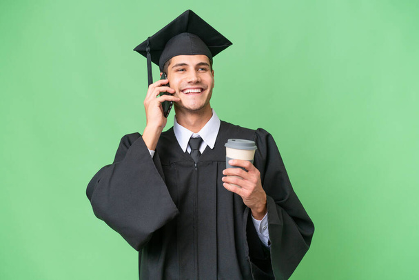 Young university graduate caucasian man over isolated background holding coffee to take away and a mobile - Photo, Image