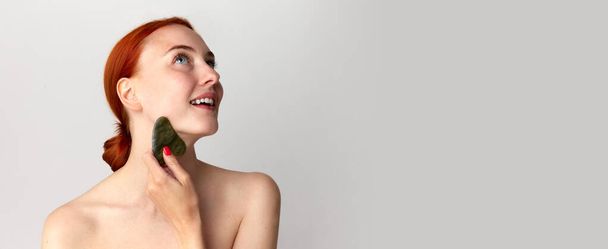 Banner with smiling, pretty girl with perfect skin and red hair doing neck massage with guasha over white studio background. Beauty, spa, cosmetology, skin care, concept. Model with well-kept skin - Photo, Image