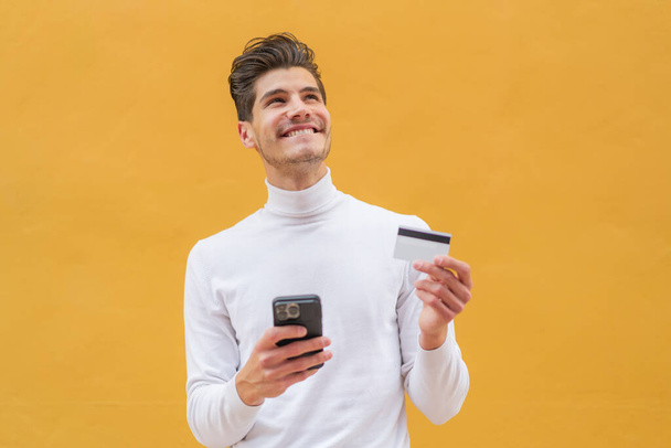 Joven hombre caucásico al aire libre comprando con el móvil con una tarjeta de crédito mientras piensa - Foto, imagen