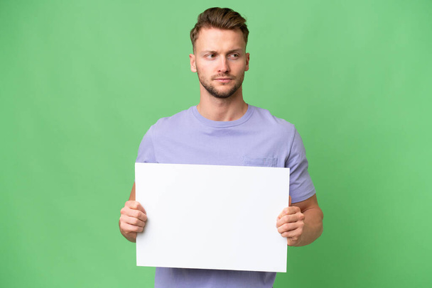 Молодой белокурый белый мужчина на изолированном фоне с пустым плакатом - Фото, изображение