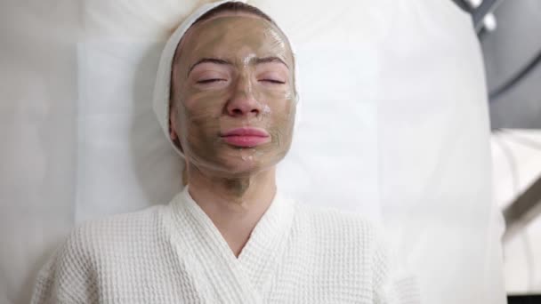 Ziemlich schöne Frau liegt in einer Schönheitsklinik, trägt ein weißes Gewand mit einer grünen Maske auf dem Gesicht zur Verjüngung der Haut. Aktive Reinigung der Haut, hilft gegen Akne, vergrößerte Poren - Filmmaterial, Video