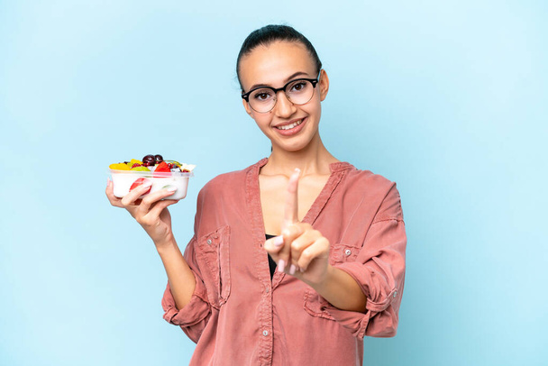 Junge arabische Frau hält eine Schale mit Früchten auf blauem Hintergrund und zeigt und hebt einen Finger - Foto, Bild