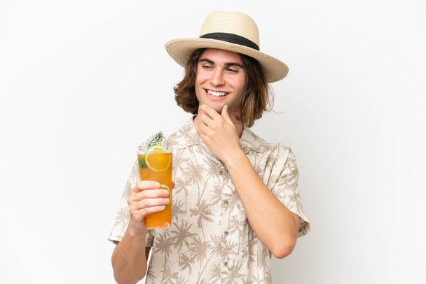 Joven hombre guapo sosteniendo un cóctel aislado sobre fondo blanco mirando hacia un lado y sonriendo - Foto, imagen
