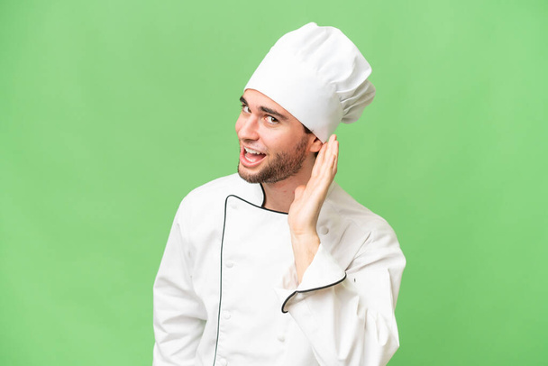 Jonge knappe chef-kok man over geïsoleerde achtergrond luisteren naar iets door de hand op het oor - Foto, afbeelding