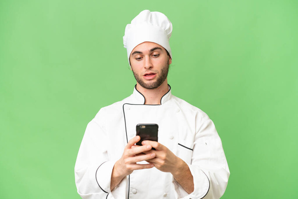 Young handsome chef man over isolated background sending a message with the mobile - Fotó, kép