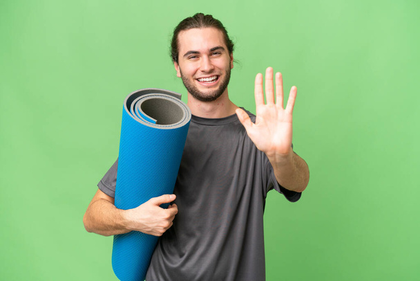 Young sport man going to yoga classes while holding a mat over isolated background counting five with fingers - Fotó, kép