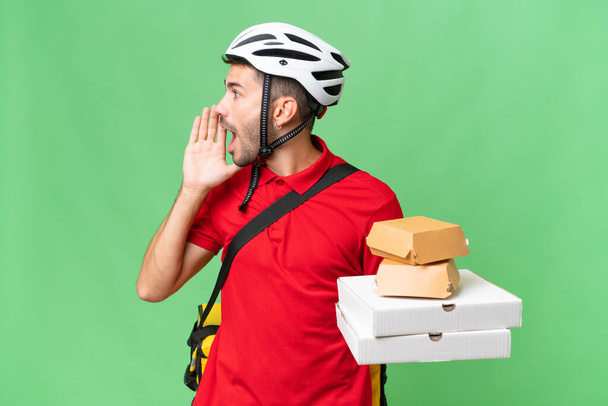 Young handsome caucasian man with thermal backpack and holding takeaway food over isolated background shouting with mouth wide open to the side - Photo, Image
