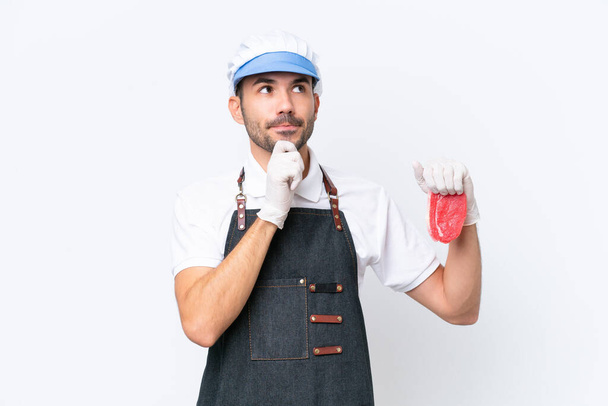 Carniceiro caucasiano homem vestindo um avental e servindo carne cortada fresca sobre fundo branco isolado com dúvidas e pensamento - Foto, Imagem