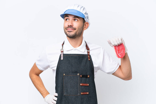 Мясник белый мужчина в фартуке и подает свежее мясо на изолированном белом фоне, позируя с руками на бедре и улыбаясь - Фото, изображение