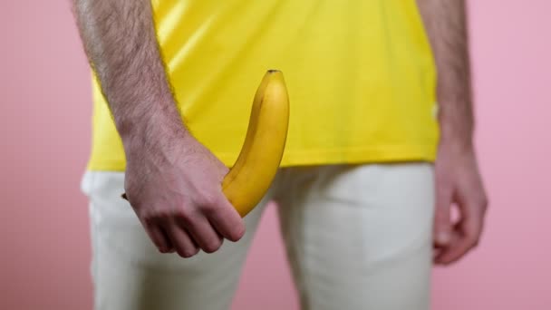 Az ember egy felemelt banánt tart a kezében a nemi szervek szintjén. Egy nő keze áthajt egy banánon. Rózsaszín háttér. A férfiak egészségének fogalma - Felvétel, videó