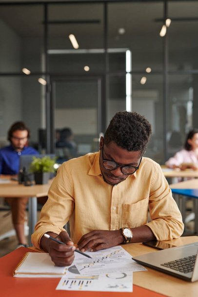 Busy young African-American businessman in eyeglasses sitting in shared workspace and analyzing chart - Valokuva, kuva