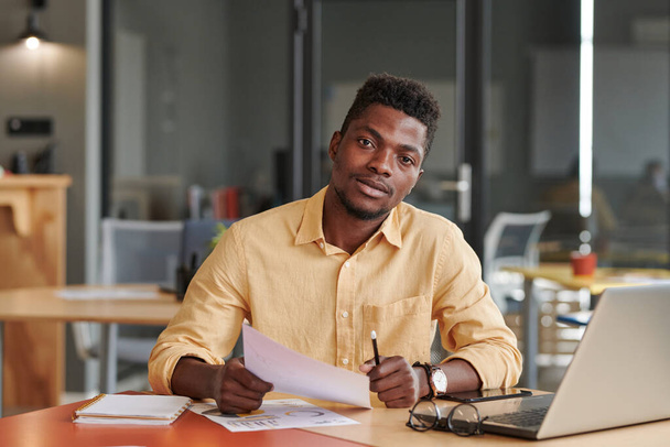 Portrait of confident young African-American office man sitting at desk with papers and laptop - Valokuva, kuva