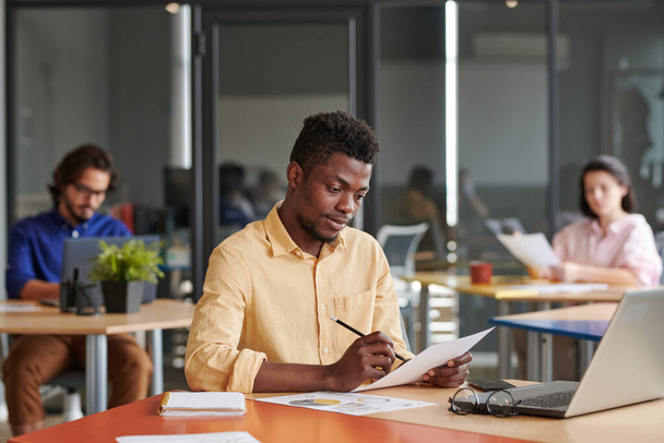 Busy young African-American business expert sitting at table and examining papers in open-space office - Foto, afbeelding